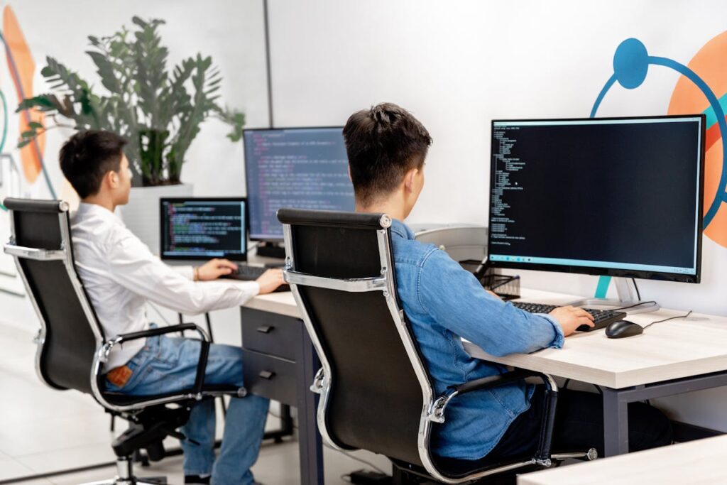 pexels-photo-7988079 Two male developers at desks programming in a modern office workspace with large monitors.